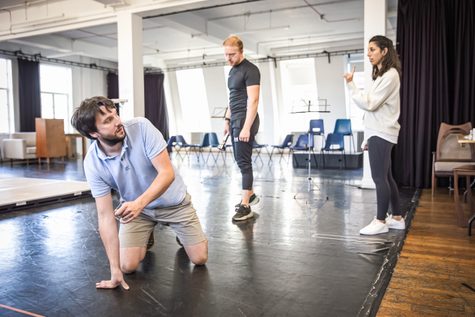 A man in a blue shirt crouches on the floor with a man and woman standing behind him