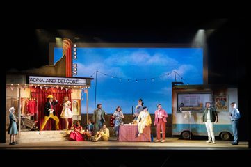 The cast of The Elixir of Love stands across the stage with a dilapidated theatre on the left and a fish and chips truck on the right.