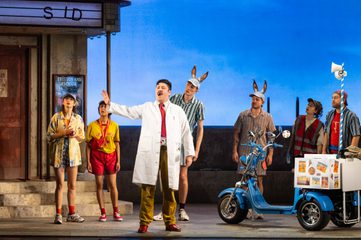 Dulcamara stands centre stage wearing a lab coat and singing to the townspeople.