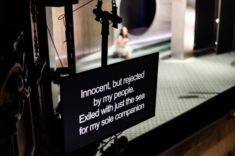 White text on a black surtitle screen displayed in front of a stage