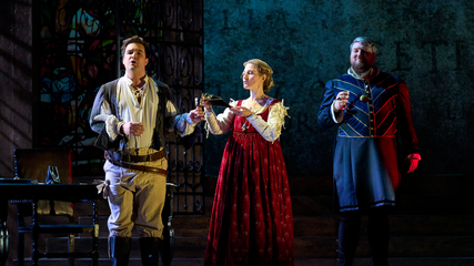 Three singers in period clothing stand on a dimly lit stage, holding gold glasses. The female pours wine into one of the cups.