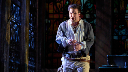 A male singer wearing period clothing stands centre stage with his hands in front of him. He looks angry.