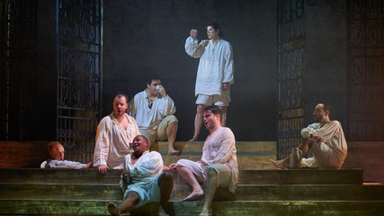 A group of male singers dressed in white pyjama-style clothing gather around steam room steps.