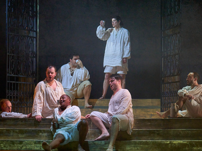 A group of male singers dressed in white pyjama-style clothing gather around steam room steps.