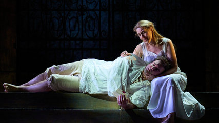 A male singer lies asleep, across the lap of a female singer who holds his head, singing. They are in white pyjama-style period clothing.