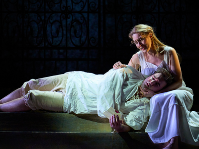 A male singer lies asleep, across the lap of a female singer who holds his head, singing. They are in white pyjama-style period clothing.