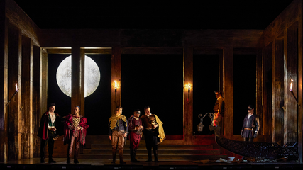 A group of male singers in period costumes stand on a dimly lit stage, with lit torches.