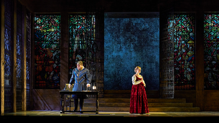 A male and female stand in period costumes on a dimly lit stage, in front of large stain glass windows. The male stands behind a table.