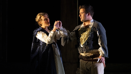 A male and female wearing period clothing stand against a black background, holding hands.