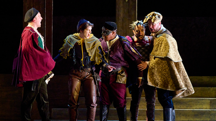 Five men wearing period clothing and masks, stand huddled centre stage and singing.