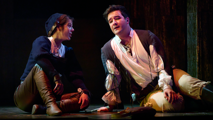 Two singers sit onstage, looking at one another and singing. They are dressed in period clothing.