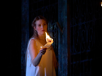Women wearing white period clothing stands holding a letter which is aflame.