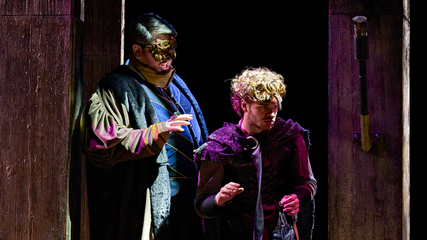 Two male singers, wearing masks and period clothing, stand suspiciously. One puts his hand out near the other's shoulder.