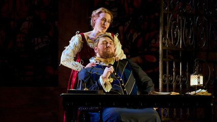 A male and female wearing period clothing are behind a table - the male is seated and the female massages his shoulders. The stage is dimly lit.