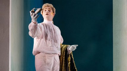 A man in a powdered wig, white shirt, breeches and gloves holds up a woman's shoe