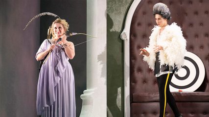 A blonde woman with a feather in her hair holds a bow as a young man in white and black struts towards her