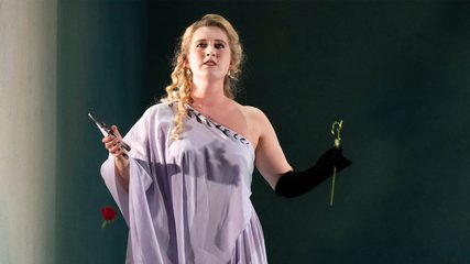 A blonde woman dressed in white stands with secateurs and a rose