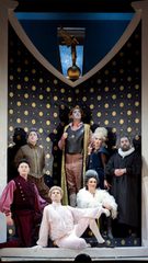Seven people stand haughtily in period costume in front of a decorative backdrop as if for a portrait