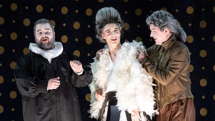 A young man in white fur and a powdered wig smiles, surrounded by a bearded man in black and a smiling man in brown jacket and breeches