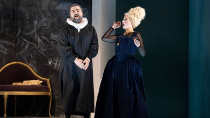 A man dressed in black and with a white ruff stands as a woman in blue with a white beehive wig looks on in shock