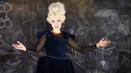 A smiling woman with a white beehive wig dressed in blue stands against a blackboard with arms outstretched