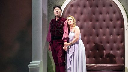 A tall man dressed in red sings in front of a neo-classical background as a blonde woman dressed in white holds his hand
