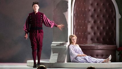 A man dressed in a red jacket and breeches stands above a blonde woman dressed in white seated on the floor