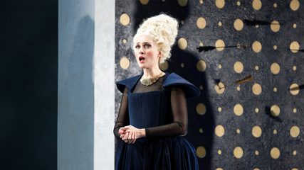 A woman with a beehive wig dressed in blue stands with her hands clasped against a background peppered with arrows