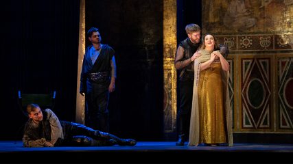 A bearded man embraces a smiling woman dressed in gold, watched by a man lying on the ground and a standing man dressed in black