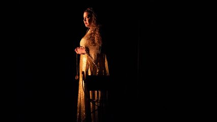 A woman with long dark hair in a gold dress stands in darkness