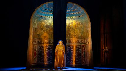 A woman in a gold dress stands in front of a wall decorated with mosaics and frescos