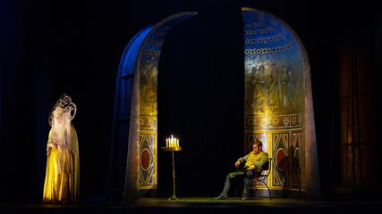 A veiled woman is threateningly observed by a seated man bathed in candlelight