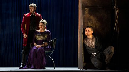A man in a red coat stands above a seated woman dressed in purple while a bearded man in dark clothing listens from behind a corner