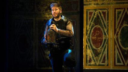 A bearded man dressed in leather armour kneels while holding a lantern