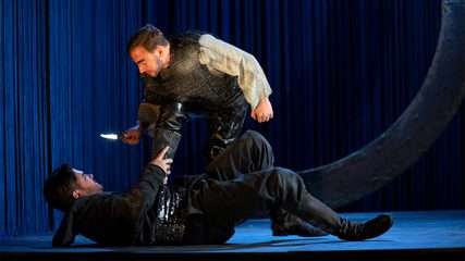 A man stands above a man in black who lies on the ground, menacing him with a dagger