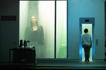 A man in white shirt and grey trousers walks into an elevator. Beside him, the figure of a woman in black leather jacket is projected on a wall.