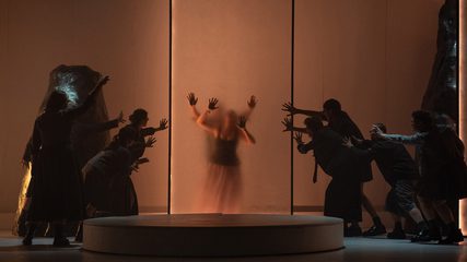 A man and woman are seen embracing through a translucent glass panel, observed by a group of silhouettes