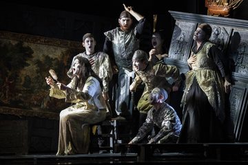 People are gathered around the Sorceress. The Sorceress sits in a chair, wearing a white dress and holding a mirror.