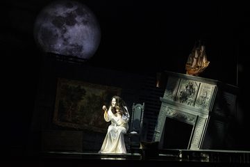 The Sorceress sits on a chair in the middle of the room. There is a moon in the left corner and a falling fireplace on the right side. The fireplace has a wooden ship above it