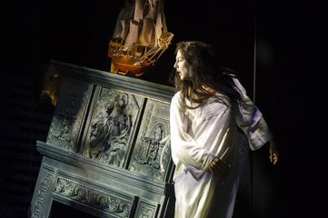 The Sorceress is wearing a full white dress and stands next to a falling fireplace