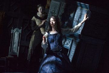 Dido sits holding onto the fireplace in the background. She holds a mirror and looks into it. The second woman is standing behind her in the shadows.