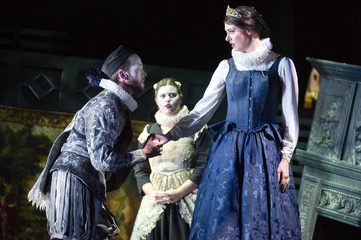 Dido stands in a black dress with a ruff. Aeneas holds her hand and is beginning to kneel. The second woman in standing in the background wearing a black and white and wearing a ruff.