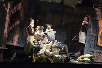 Four people sit whilst a man lies on the floor. A man is standing in the corner. The background is all wooden and carved