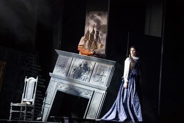 There is a chair and a fireplace that is falling. Above the fire place is a ship. Dido stands next to the fire place in a black dress.