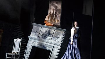 There is a chair and a fireplace that is falling. Above the fire place is a ship. Dido stands next to the fire place in a black dress.