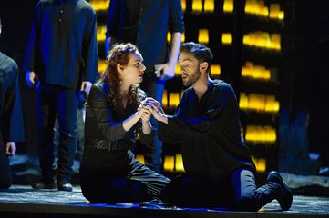 Dido and Aeneas sit on the floor holding hands. There are people in the background and a wall of lanterns.