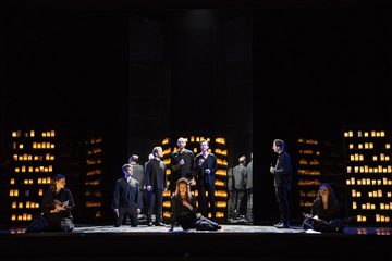 Dido, Belinda and Second Woman sit along the front. Men stand in the background. The walls are filled with lit lanterns