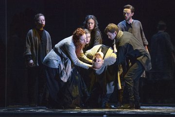 A man is being held up by four people, staring into the audience.
