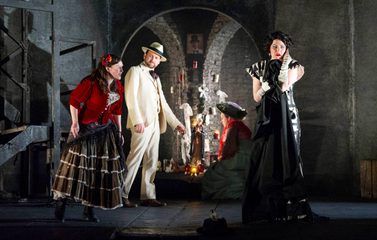 Zerlina and Donna Elvira are at the front of the stage. Zerlina is shouting at Donna Elvira and Donna Elvira has her hands over her ears. Don Ottavio watches from behind