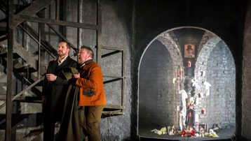 Don Giovanni and Leporello are standing stage right next to a staircase. Leporello is holding his coat and Don Giovanni.